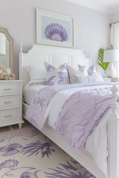 a white bed topped with purple pillows next to a dresser and mirror in a bedroom