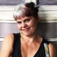 a woman with grey hair wearing a black top and smiling at the camera while leaning against a wall