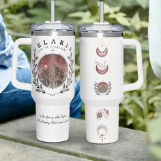 two white travel mugs sitting next to each other on a wooden table with trees in the background