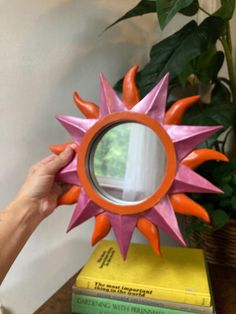 a person is holding up a pink and orange starburst shaped mirror in front of a stack of books