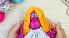 a hand holding a small doll with yellow hair and blue eyes in front of yarn balls