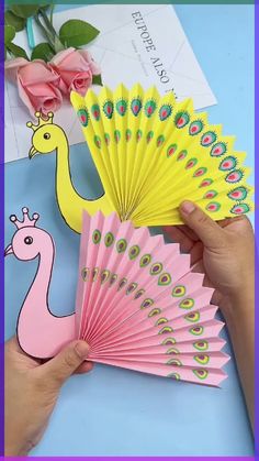 a hand holding a pink and yellow paper fan next to a flower on a blue table