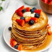 a stack of pancakes topped with fruit on top of a white plate next to a bowl of strawberries and blueberries