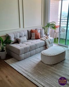 a living room with a couch, ottoman and potted plant on the floor in front of large windows