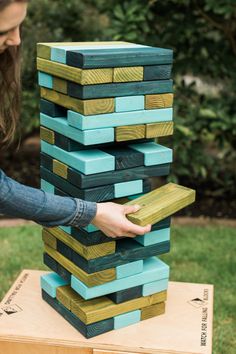 a woman is placing blocks on top of a wooden block tower in front of some grass