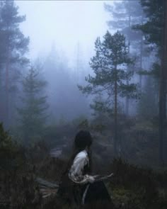 a woman sitting in the middle of a forest on a foggy day with her back to the camera