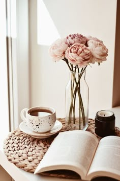 a table topped with a vase filled with flowers and a cup of coffee next to an open book