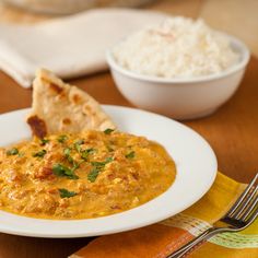 a white plate topped with curry next to a bowl of rice and pita bread