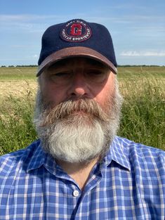 a man with a beard wearing a hat in the middle of a grassy field on a sunny day