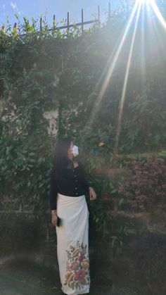 a woman standing in front of a stone wall drinking from a cup with the sun shining behind her