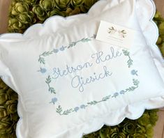 a white pillow with embroidered words on it sitting in front of some green plants and pine cones