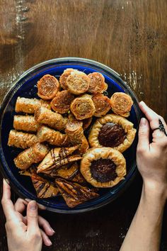 two hands reaching for pastries in a blue plate on a wooden table with wood grain