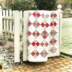 a quilt hanging on the side of a white picket fence in front of a garden