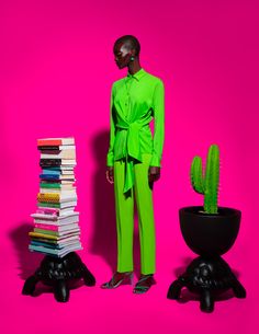 a mannequin dressed in green stands next to a stack of books and a cactus