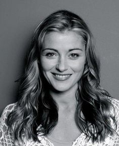 a black and white photo of a woman with long hair smiling at the camera, in front of a gray background