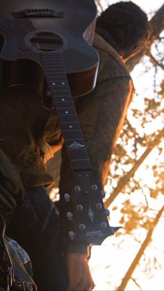 a man holding a guitar in his right hand and looking down at the ground with trees behind him