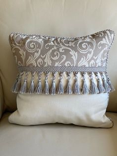 a white and gray pillow with tassels on it sitting on a couch in front of a wall