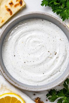 a bowl filled with white sauce next to sliced lemons and pita bread