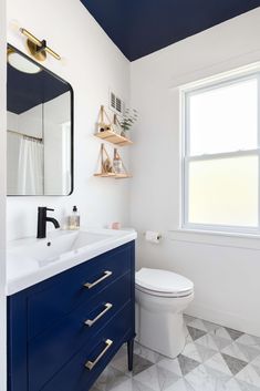 a white toilet sitting next to a blue cabinet in a bathroom under a window with wooden shelves