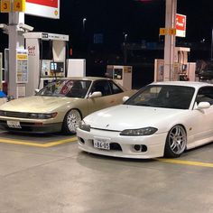 two white cars parked in front of gas pumps at night, one is gold and the other is silver