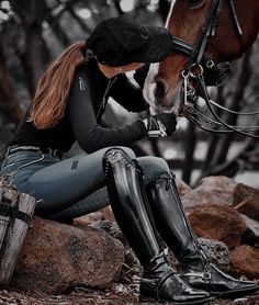 a woman in black riding boots sitting on rocks next to a horse