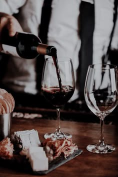 a man pouring wine into two glasses on top of a wooden table with cheese and crackers