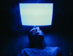 a man laying down in the dark with his head up on a monitor screen that is glowing blue