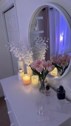 flowers and candles on a white table in front of a round mirror