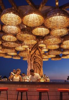 a man and woman sitting at a bar under wicker lamps