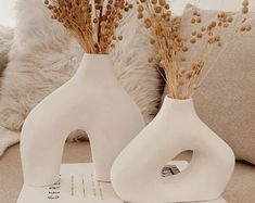 two white vases sitting on top of a table next to each other with dried plants in them