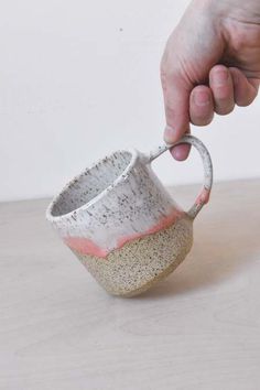 a hand is holding a small cup with a handle on the table next to a white wall