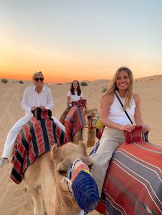 three people riding camels in the desert at sunset