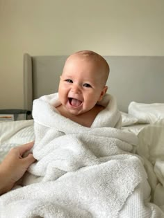 a smiling baby wrapped in a towel on top of a bed with someone's hand