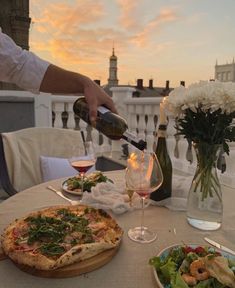 a person pouring wine into a glass near a pizza and salad on a table outside