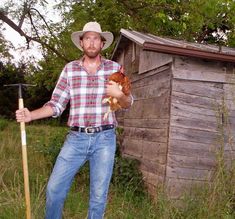 a man holding a chicken in his right hand while standing next to a wooden shack