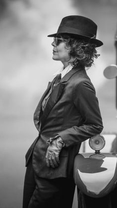 a woman in a suit and hat standing next to a car with her hand on the door handle