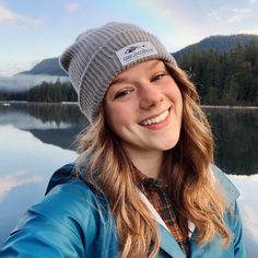 a woman is taking a selfie in front of a lake with mountains and trees