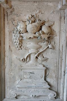 a white vase with fruit and flowers on it in front of a stone wall panel