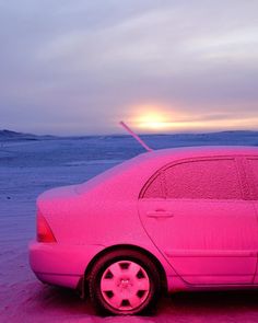 a pink car is parked in the snow at sunset or dawn, with the sun setting behind it