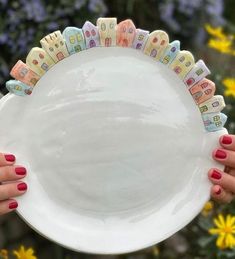 two hands holding a white plate with houses painted on the side and yellow flowers in the background