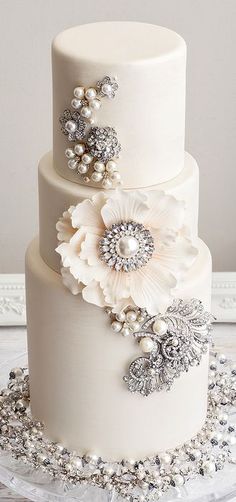 a three tiered wedding cake with white flowers and pearls on the top, sitting on a glass platter