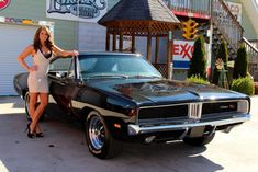 a woman standing next to a black muscle car in front of a building with a sign on it