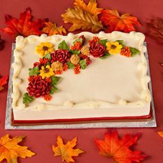 a cake decorated with flowers and leaves on top of a red table next to autumn leaves