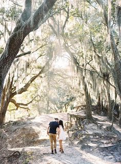 a man and woman walking down a dirt path in front of trees with spanish moss