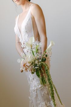 a woman in a white wedding dress holding a bouquet of flowers and greenery with her hands behind her back