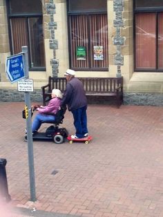 an elderly man pushing a woman on a motorized scooter down the street in front of a building