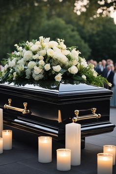 a casket with white flowers and candles on the ground in front of it, surrounded by people