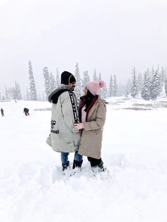 two people standing in the snow with trees in the background and one person wearing a pink hat