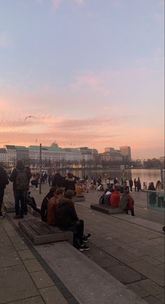 many people are sitting on benches near the water at sunset or dawn, with buildings in the background