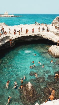 many people are swimming in the clear blue water near an arch shaped bridge that is built into the rocks